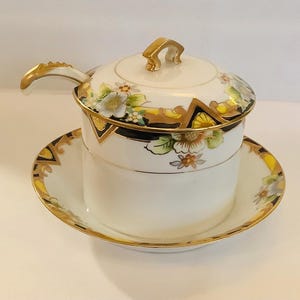 May include: A white ceramic sugar bowl with a lid and a small spoon, set on a matching saucer. The bowl and saucer are decorated with floral patterns, gold trim, and black accents. The handle and lid finial are gold.