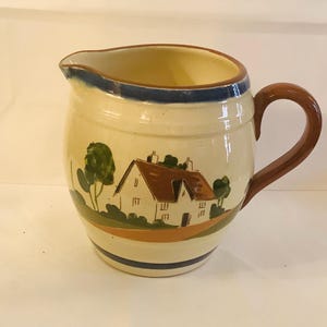 May include: A cream-colored ceramic pitcher with a brown handle. The pitcher has a blue rim and a painted scene of a white cottage with brown roof and green trees.