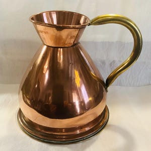May include: A polished copper pitcher with a wide, flared rim and a rounded body. The vessel features a curved brass handle attached to the side, providing a contrast in color and material. The base is circular and flat, supporting the pitcher.