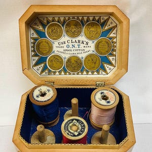 May include: Wooden sewing box with a hinged lid, featuring spools of blue, pink, and red thread. The interior is lined with blue fabric. The lid displays vintage graphics and text, including "CLARK'S O.N.T." and various medallions.