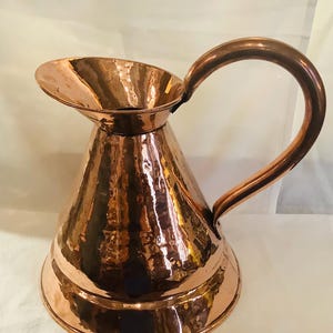 May include: A polished copper pitcher with a wide, flared base and a conical body. The surface features a distinct hammered texture throughout. It includes a curved handle attached to the side and a wide, open spout for pouring liquids.
