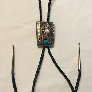 May include: A silver bolo tie with a rectangular pendant featuring a floral design and turquoise and coral stones. The tie is made of black braided leather.