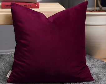 May include: A deep burgundy velvet pillow with a square shape. The pillow is sitting on a gray rug in front of a wooden table with books stacked on top.