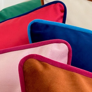 May include: A stack of colorful throw pillows with contrasting piping. The pillows are made of velvet and feature a variety of colors, including pink, blue, red, green, and brown.