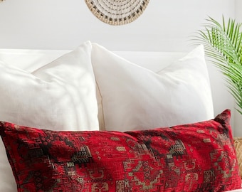Puede incluir: Una cama blanca con almohadas blancas y un cojín lumbar de patrón rojo. La cama está en un dormitorio con paredes blancas y una planta en una cesta tejida.