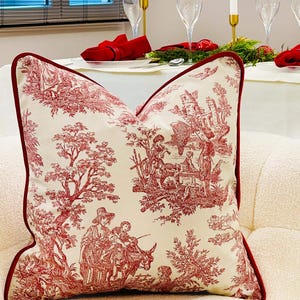 Op de afbeelding: Crèmekleurig decoratief kussen met een rood toile de Jouy-patroon met figuren, bomen en gebouwen. Het kussen heeft een rode rand en ligt op een witte bank. Op de achtergrond is een eettafel te zien die is gedekt voor een formele gelegenheid.