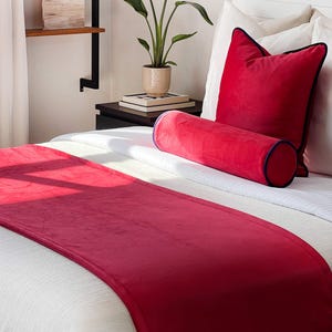 May include: A scarlet red bed runner, rectangular pillow, and bolster pillow on a white bed. The rectangular pillow has a black trim. The bolster pillow has a blue trim. The text "Scarlet Red" is in the bottom left corner.