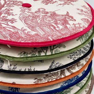 May include: A stack of round, decorative cushions with a toile pattern in red and white. Each cushion has a different colored trim, including pink, green, black, orange, blue, and brown. A red button is visible on the top cushion.