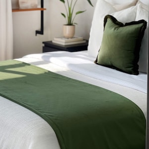 May include: A moss green bed runner on a white bed, with a matching green pillow with a dark trim. The bed has white sheets and pillows. A potted plant and a shelf are in the background.