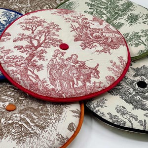 May include: A collection of round cushions with toile patterns in various colors. The cushions feature scenes of people and landscapes in shades of blue, red, brown, green, and black, with contrasting colored piping and a central button.