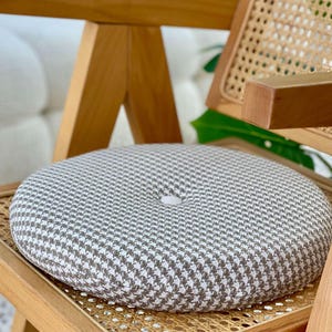 May include: A circular seat cushion with a grey and white houndstooth pattern rests on a wooden chair with a woven cane back. The cushion has a central button. The chair has a light wood finish.