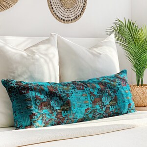 May include: A long, rectangular turquoise cushion with a geometric pattern in brown and gold tones sits on a white bed. White pillows and a woven wall hanging are visible in the background, creating a cozy atmosphere.