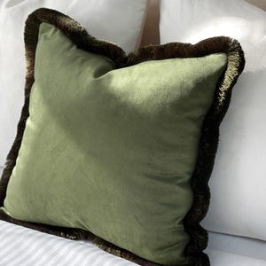 May include: A square, olive green pillow with a dark brown fringe trim. The pillow is made of a soft, velvety material and is placed on a bed with white pillows in the background. The pillow is a decorative item.