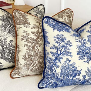 K&ouml;nnte beinhalten: Drei dekorative Zierkissen mit Toile-de-Jouy-Mustern. Die Kissen haben schwarze, braune und blaue Paspeln und zeigen Szenen von Menschen und B&auml;umen im klassischen Toile-Stil. Die Kissen liegen auf einem wei&szlig;en Sofa.