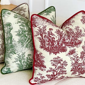 K&ouml;nnte beinhalten: Vier dekorative Kissen mit Toile-de-Jouy-Muster. Die Kissen haben einen cremefarbenen Hintergrund mit Szenen in Braun, Gr&uuml;n und Rot. Jedes Kissen hat eine kontrastierende Bord&uuml;re in Braun, Gr&uuml;n bzw. Rot. Die Kissen sind auf einem wei&szlig;en Sofa angeordnet.