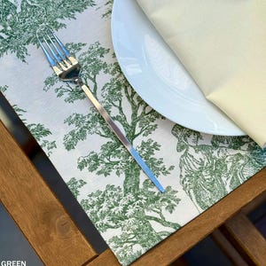 Pode incluir: Um lugar americano retangular com um padrão toile verde e branco de árvores e figuras, com um garfo de aço inoxidável e um guardanapo cor de creme dobrado sobre um prato branco, tudo sobre uma superfície de madeira com a palavra GREEN.