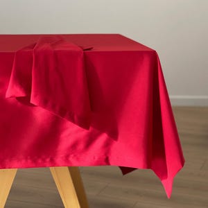 Può includere: Una tovaglia rosso vivo drappeggiata su un tavolo di legno. Il tessuto è liscio e sembra essere fatto di un materiale morbido. Un tovagliolo rosso abbinato è piegato e posto sul tavolo. Il tavolo è posto su uno sfondo bianco.