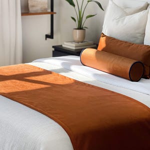 Könnte beinhalten: Ein Bett mit weißer Bettwäsche, mit einem rostfarbenen Bettläufer und passenden Kissen. Ein zylindrisches Nackenkissen mit marineblauer Paspelierung ist ebenfalls vorhanden. Die Szene wird von natürlichem Licht beleuchtet.