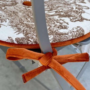 May include: A round chair cushion with a brown and white toile pattern, trimmed with orange velvet. The cushion is secured to the chair with an orange velvet tie.