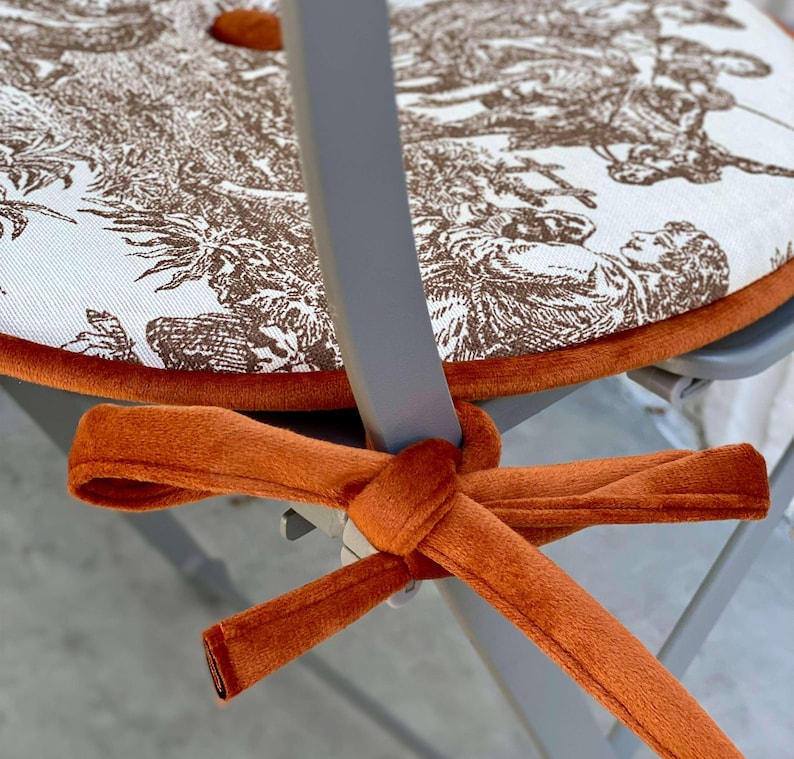 May include: A round chair cushion with a brown and white toile pattern, trimmed with orange velvet. The cushion is secured to the chair with an orange velvet tie.