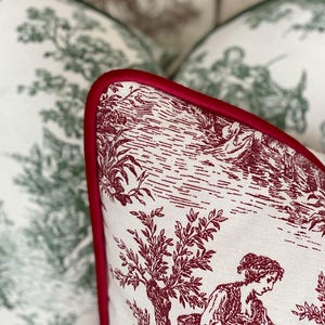 K&ouml;nnte beinhalten: Nahaufnahme eines dekorativen Kissens mit einem rot-wei&szlig;en Toile-Muster. Das Kissen hat einen roten Rand und zeigt eine l&auml;ndliche Szene mit Figuren, B&auml;umen und Geb&auml;uden. Im Hintergrund sind weitere Kissen mit einem gr&uuml;n-wei&szlig;en Muster zu sehen.
