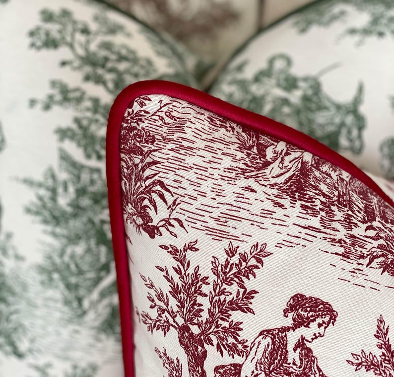 K&ouml;nnte beinhalten: Nahaufnahme eines dekorativen Kissens mit einem rot-wei&szlig;en Toile-Muster. Das Kissen hat einen roten Rand und zeigt eine l&auml;ndliche Szene mit Figuren, B&auml;umen und Geb&auml;uden. Im Hintergrund sind weitere Kissen mit einem gr&uuml;n-wei&szlig;en Muster zu sehen.