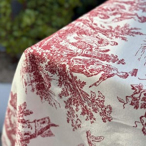 Op de afbeelding: Een wit stoffen tafelkleed met een herhalend rood toile de jouy patroon. Het ontwerp toont gedetailleerde scènes van figuren in buitenomgevingen, omringd door bomen, gebladerte en landschapselementen gedrukt in dieprode inkt op een witte ondergrond.