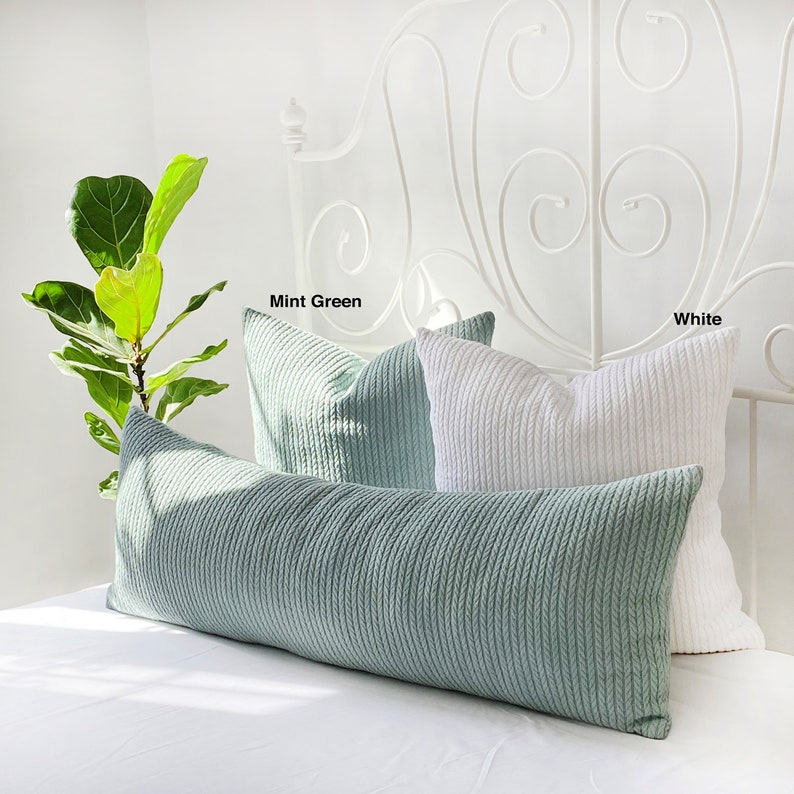 May include: Two decorative pillows on a white bed. The larger pillow is a mint green body pillow with a cable knit texture. The smaller pillow is white with a cable knit texture. The text "Mint Green" and "White" are written above the pillows.