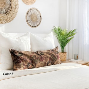 May include: A long, rectangular cushion with a geometric pattern in shades of brown, red, and green. The cushion is placed on a white bed with white pillows and woven wall hangings in the background. The text "Color 3" is visible.