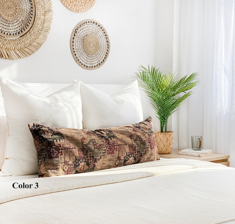 May include: A long, rectangular cushion with a geometric pattern in shades of brown, red, and green. The cushion is placed on a white bed with white pillows and woven wall hangings in the background. The text "Color 3" is visible.