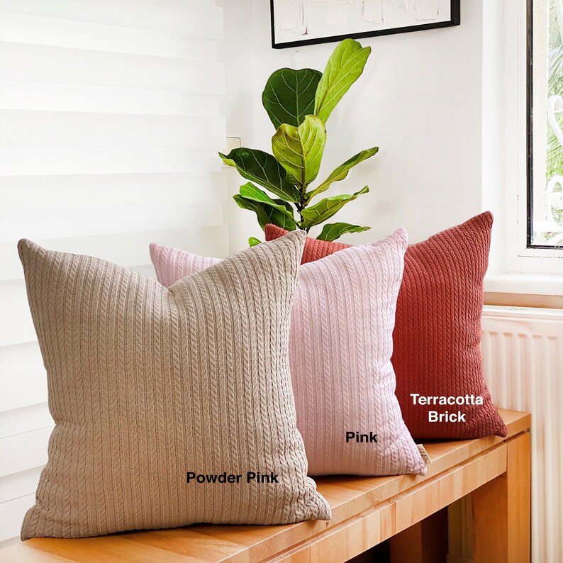 May include: Three decorative throw pillows with a cable knit texture. The pillows are in shades of powder pink, pink, and terracotta brick.
