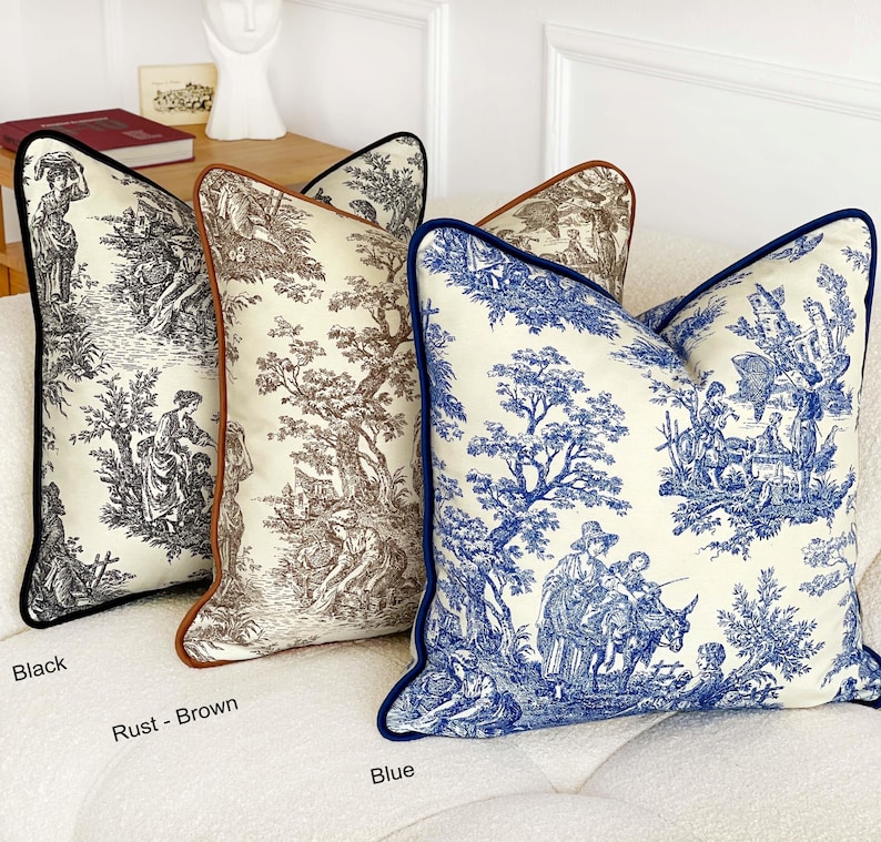 May include: Decorative throw pillows with toile scenes in black, rust-brown, and blue. Each pillow features a contrasting border: black, rust-brown, and blue. The pillows are on a white sofa. The pillows have a classic, elegant design.