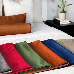 Peut inclure: Un lit est fait avec des oreillers blancs et une couette blanche. Plusieurs coussins d&eacute;coratifs en velours color&eacute;s et du tissu pli&eacute; sont expos&eacute;s. Les couleurs incluent le rouge, le vert, l'orange, le bleu et le beige. Une plante en pot est pos&eacute;e sur une table de chevet noire.