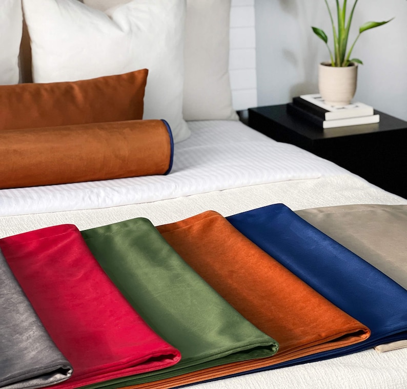 Peut inclure: Un lit est fait avec des oreillers blancs et une couette blanche. Plusieurs coussins d&eacute;coratifs en velours color&eacute;s et du tissu pli&eacute; sont expos&eacute;s. Les couleurs incluent le rouge, le vert, l'orange, le bleu et le beige. Une plante en pot est pos&eacute;e sur une table de chevet noire.