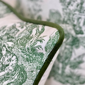 May include: Close-up of a decorative pillow with a green and white toile pattern. The pillow features a detailed pastoral scene and is trimmed with a dark green velvet piping.