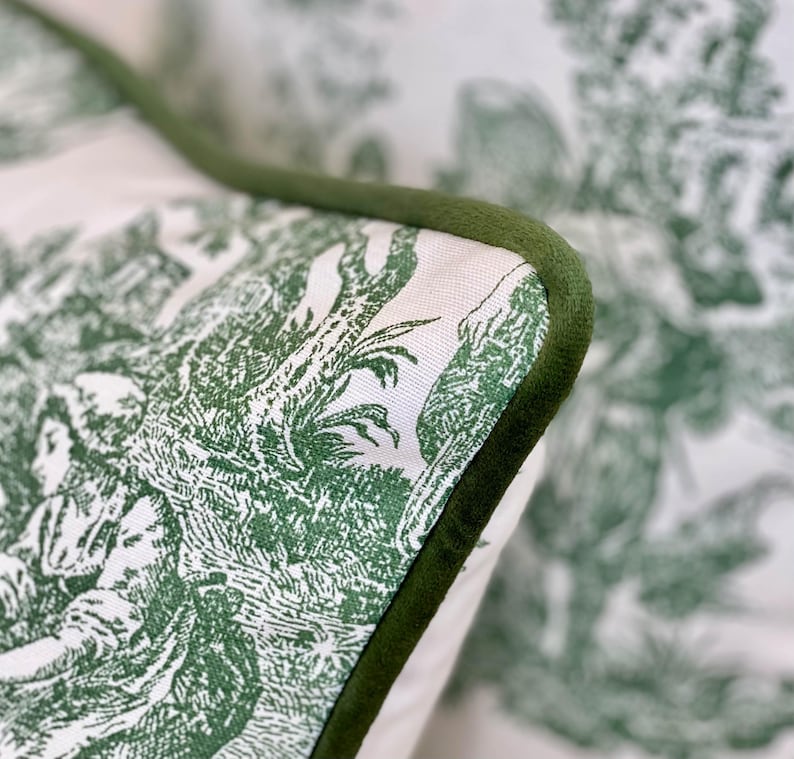 May include: Close-up of a decorative pillow with a green and white toile pattern. The pillow features a detailed pastoral scene and is trimmed with a dark green velvet piping.