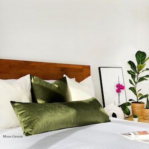May include: A bed with white bedding and a green velvet lumbar pillow. There are two white square pillows and one green velvet square pillow on the bed. The headboard is made of wood. There is a framed picture with a pink orchid on the wall behind the bed. A potted plant is on the nightstand next to the bed. Moss Green.