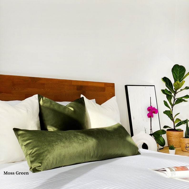 May include: A bed with white bedding and a green velvet lumbar pillow. There are two white square pillows and one green velvet square pillow on the bed. The headboard is made of wood. There is a framed picture with a pink orchid on the wall behind the bed. A potted plant is on the nightstand next to the bed. Moss Green.