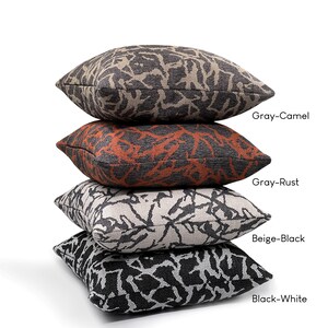 May include: Four decorative throw pillows stacked on top of each other. The pillows are made of a textured fabric with a black and white abstract pattern. The top pillow is gray with a camel colored pattern, the second pillow is gray with a rust colored pattern, the third pillow is beige with a black pattern, and the bottom pillow is black with a white pattern. The pillows are labeled with their color combinations: Gray-Camel, Gray-Rust, Beige-Black, and Black-White.