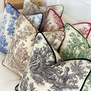May include: A collection of decorative throw pillows with various toile patterns in blue, brown, green, and red. Each pillow features a contrasting trim in colors like navy, brown, and black. The pillows are arranged on a white surface.