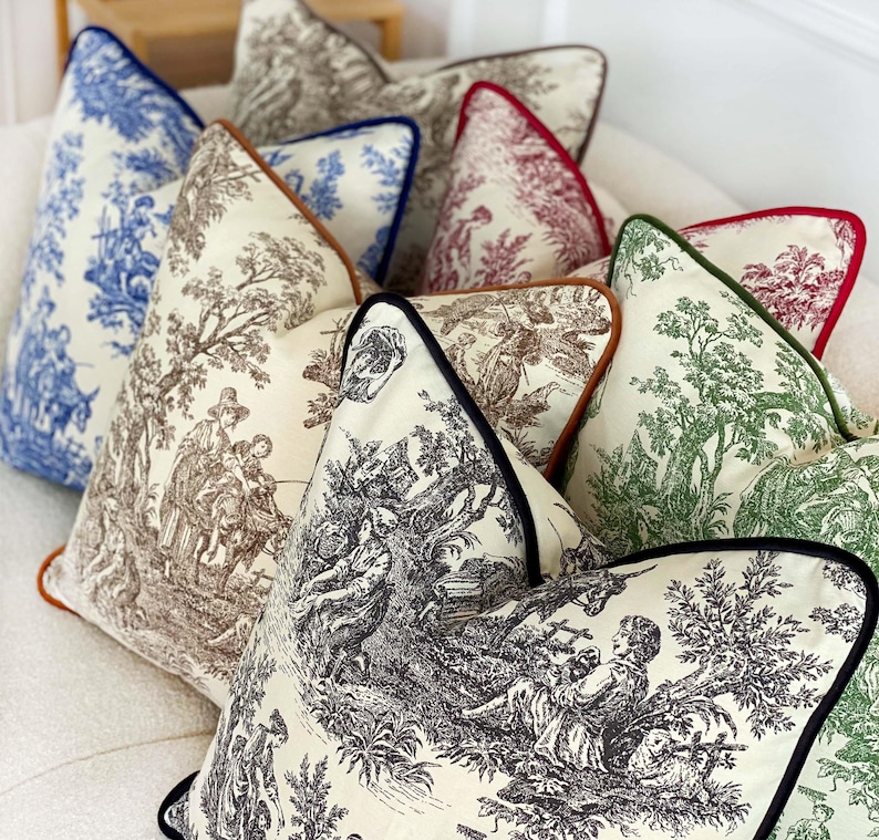 May include: A collection of decorative throw pillows with various toile patterns in blue, brown, green, and red. Each pillow features a contrasting trim in colors like navy, brown, and black. The pillows are arranged on a white surface.