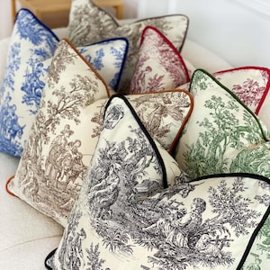 K&ouml;nnte beinhalten: Eine Sammlung dekorativer Kissen mit verschiedenen Toile-de-Jouy-Mustern in Blau, Braun, Gr&uuml;n und Rot. Jedes Kissen hat eine kontrastierende Paspel in Marineblau, Braun und Schwarz. Die Kissen sind auf einer wei&szlig;en Oberfl&auml;che angeordnet.