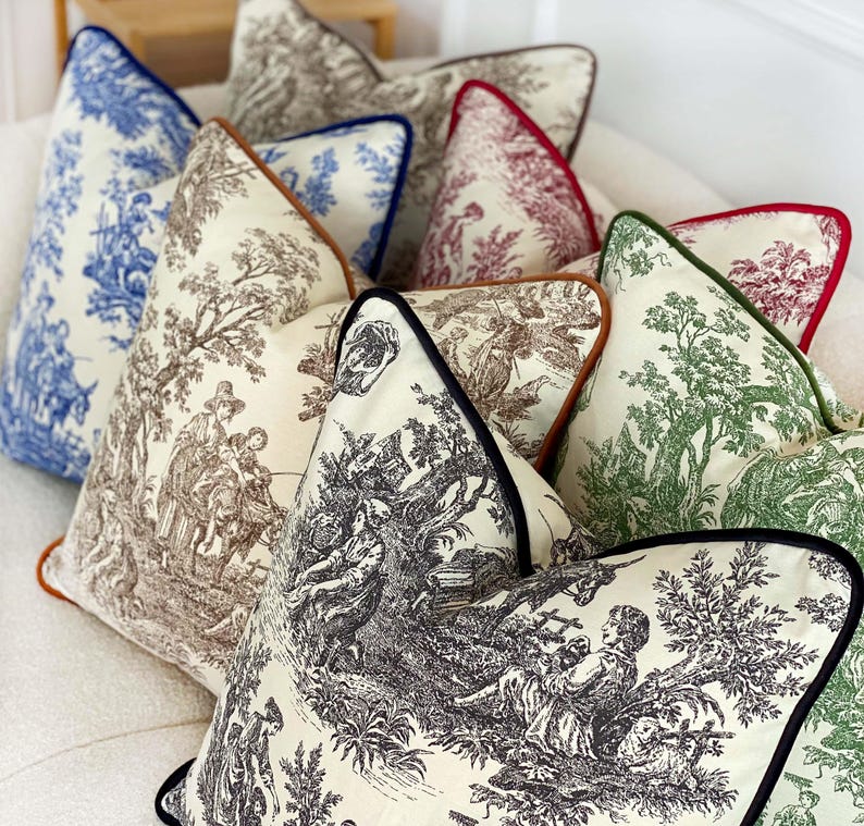 K&ouml;nnte beinhalten: Eine Sammlung dekorativer Kissen mit verschiedenen Toile-de-Jouy-Mustern in Blau, Braun, Gr&uuml;n und Rot. Jedes Kissen hat eine kontrastierende Paspel in Marineblau, Braun und Schwarz. Die Kissen sind auf einer wei&szlig;en Oberfl&auml;che angeordnet.