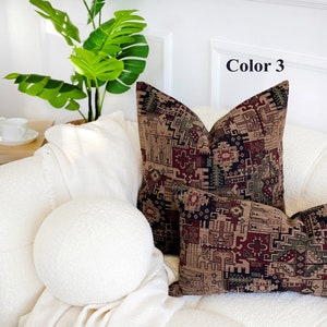 May include: Two decorative throw pillows with a geometric pattern in shades of brown, red, green, and black. The pillows are on a white couch with a white throw blanket and a round white pillow.