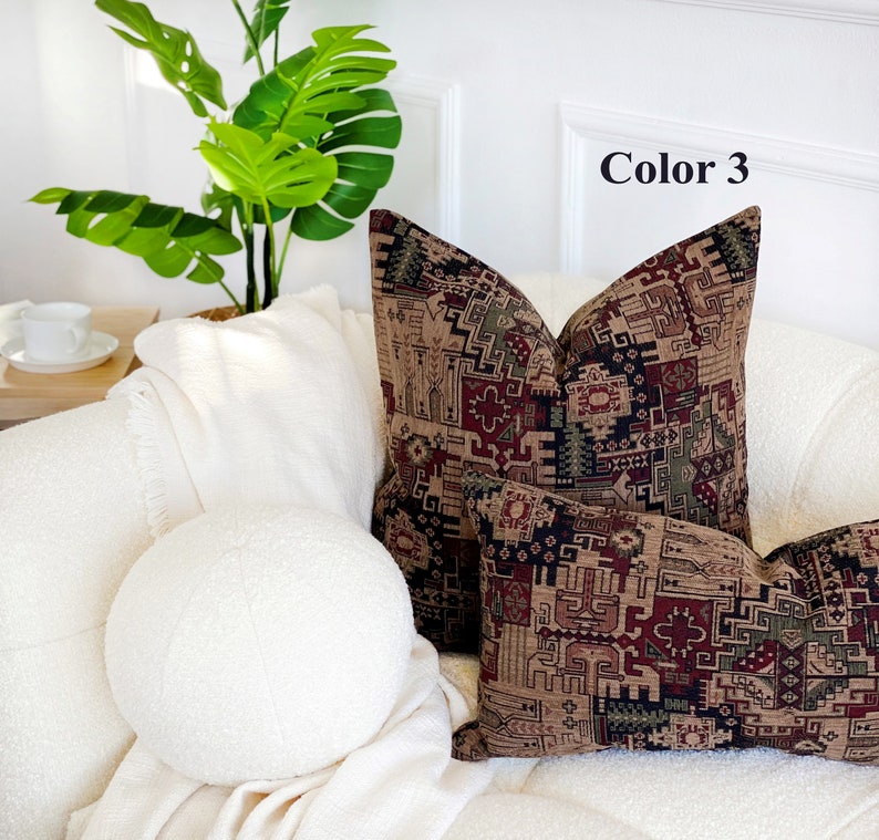 May include: Two decorative throw pillows with a geometric pattern in shades of brown, red, green, and black. The pillows are on a white couch with a white throw blanket and a round white pillow.