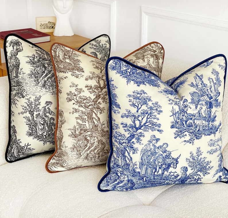 K&ouml;nnte beinhalten: Drei dekorative Zierkissen mit Toile-de-Jouy-Mustern. Die Kissen haben schwarze, braune und blaue Paspeln und zeigen Szenen von Menschen und B&auml;umen im klassischen Toile-Stil. Die Kissen liegen auf einem wei&szlig;en Sofa.