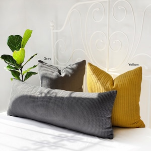 May include: Two decorative pillows, one gray and one yellow, with a cable knit pattern. The pillows are on a white bed with a white metal headboard.