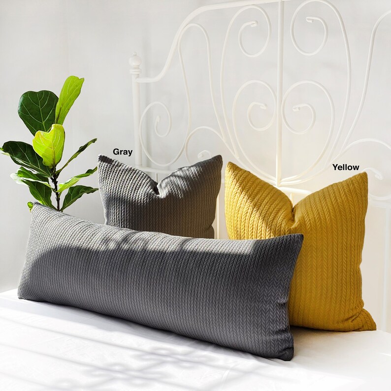 May include: Two decorative pillows, one gray and one yellow, with a cable knit pattern. The pillows are on a white bed with a white metal headboard.