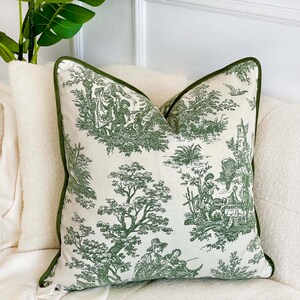 May include: Decorative throw pillow with a green and white toile pattern featuring pastoral scenes. The pillow has a green border and is placed on a white surface. The pillow is square-shaped and adds a touch of elegance to any room.