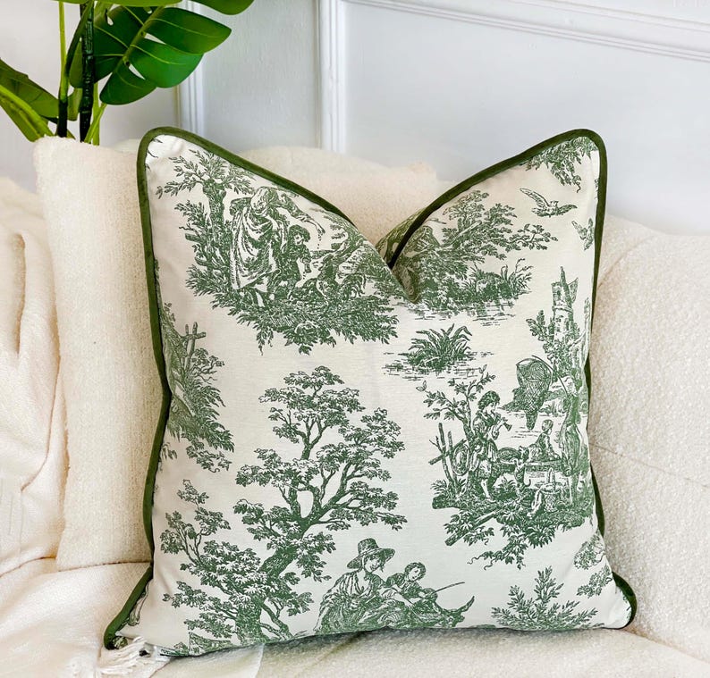 May include: Decorative throw pillow with a green and white toile pattern featuring pastoral scenes. The pillow has a green border and is placed on a white surface. The pillow is square-shaped and adds a touch of elegance to any room.